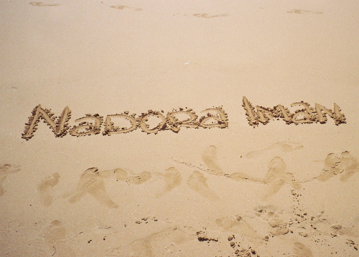 The name 'Nadora Imam' written in the sand on a beach, with footprints and shell-like decorations scattered around it.