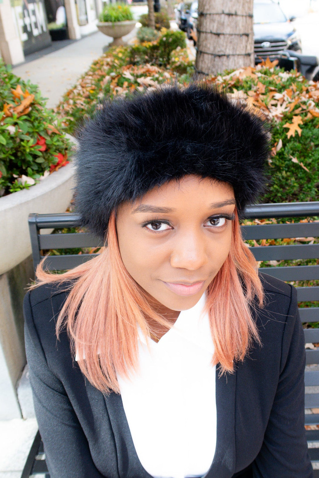Woman wearing a black fur hat and black blazer sitting on a bench outdoors.