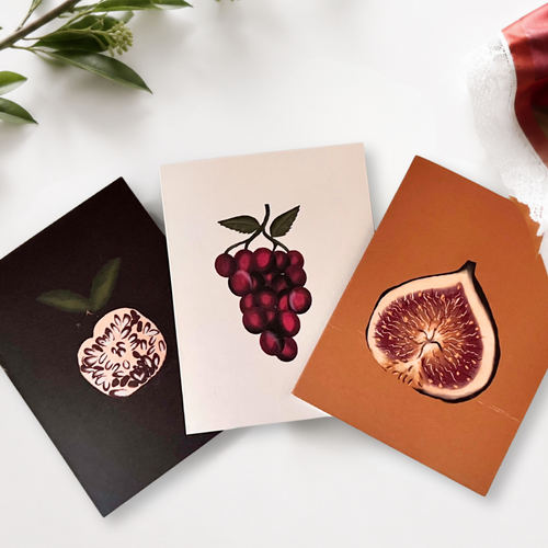 Three botanical fruit art prints arranged on a white surface with decorative greenery and a red fabric prop. The prints depict a pomegranate, a cluster of grapes, and a fig slice, each with a detailed cross-section illustration.